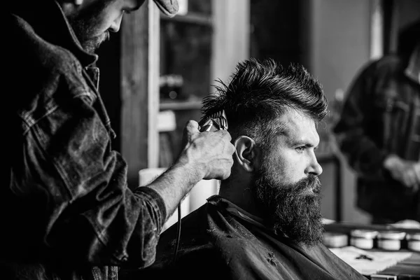 Barbier avec tondeuse à cheveux travaille sur la coiffure pour barbe homme fond de salon de coiffure. Barbier coiffant les cheveux d'un client barbu brutal avec tondeuse. Concept de mode de vie hippie. Client Hipster se faire couper les cheveux — Photo