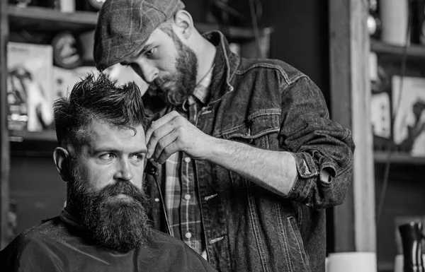 Fryzjer w kurtka dżinsowa zajęty z przycinania hipster, barbershop tła. Klient z broda i wąsy pokryta z cape. Praca Fryzjer na fryzury z włosów clipper sprzęt. Koncepcja tworzenia obrazu — Zdjęcie stockowe