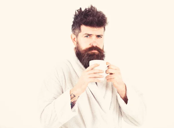 Macho drowsy, sleepy with strict face drinks coffee in morning. Morning rituals concept. Man with beard and disheveled hair stands in bathrobe, holds mug with tea or coffee, white background