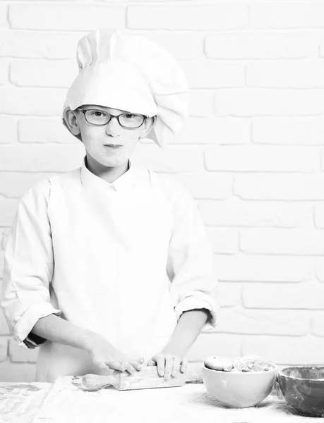 Young boy cute cook chef in white uniform and hat on stained face flour with glasses cooking on table with rolling pin and colorful bowls with chocolate cookies on brick wall background — Stock Photo, Image