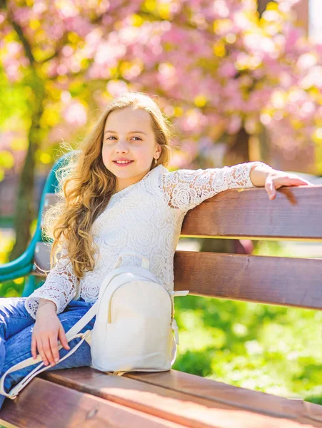 Dziewczyna na uśmiechniętą twarz siedzi na ławce, sakura na rozmytym tłem. Słodkie dziecko z plecaka cieszyć się słoneczny wiosenny dzień. Koncepcja akcesoria mody. Relaks podczas spaceru w parku, w pobliżu cherry blossom Girl — Zdjęcie stockowe