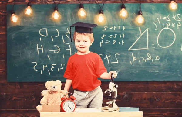 Niño, pupila sonriente cerca del microscopio. Primero interesado en estudiar, educación. Un concepto extravagante. Niño en gorra de graduado cerca del microscopio en el aula, pizarra en el fondo — Foto de Stock