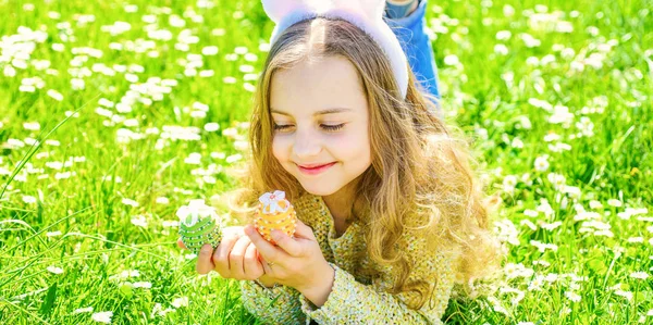 Child with cute bunny ears lying at meadow. Little girl hunting for Easter egg in spring garden on Easter day, traditional celebration. Cute child lay on grass with egg in hands. Easter feast concept