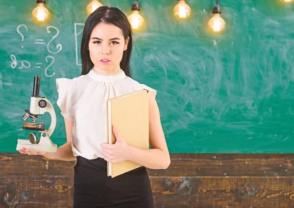 Conceito de biologia. Professor de biologia detém livro e microscópio. Senhora em desgaste formal no rosto calmo na sala de aula. Lady cientista detém livro e microscópio, quadro em segundo plano, espaço de cópia — Fotografia de Stock