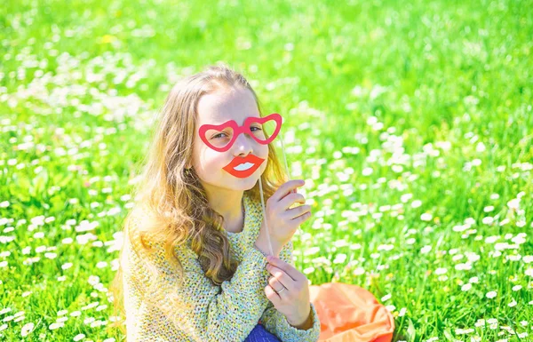 Love from first sight concept. Girl on happy face spend leisure outdoors. Girl sits on grass at grassplot, green background. Child posing with cardboard heart shaped eyeglasses and smiling mouth