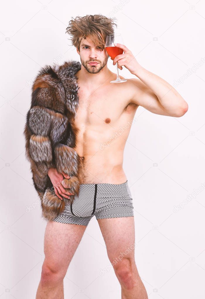 Sexy sleepy macho tousled hair drink wine or alcohol isolated on white. Luxury lifestyle and wellbeing. Richness and luxury concept. Guy attractive posing fur coat on naked body. Luxury status symbol