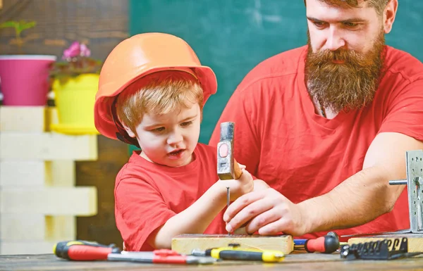 Little assistant concept. Father with beard teaching little son to use tools, hammering, chalkboard on background. Boy, child busy in protective helmet learning to use hammer with dad