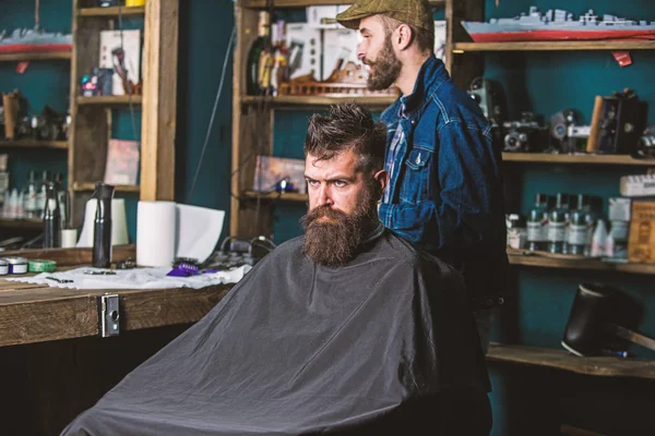 Un cliente hipster che si taglia i capelli. Cliente con barba pronta per la rifilatura o toelettatura. Uomo con la barba coperta di mantello nero in attesa mentre il barbiere cambia grado clipper. Concetto di processo taglio capelli — Foto Stock