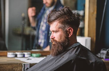 Siyah pelerin ile kaplı sakallı salonların sandalye, ayna arka plan oturur. Sakallı hippi Kuaför ve saç kesimi için bekler. Hipster berber sakal müşterilerinden olan adam. Berber kavramı
