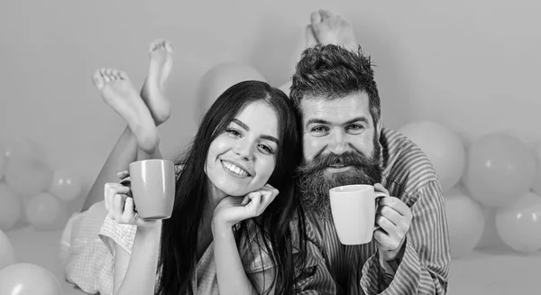 Homem e mulher em rostos sorridentes jazem, fundo rosa. Casal apaixonado bebe café na cama. Casal relaxar de manhã com café. Conceito de tradição familiar. Homem e mulher de roupas domésticas, pijamas — Fotografia de Stock