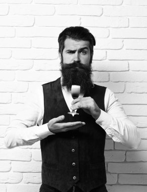 Uzun sakallı ve bıyıklı yakışıklı adamın ciddi yüzünde şık saçlar var. Beyaz tuğla duvar arka planında klasik süet deri yelek giymiş alkolik bardakları tutuyor.