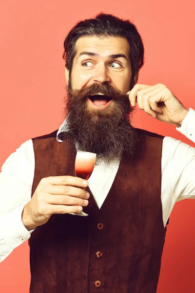 Όμορφος γενειοφόρος άνδρας με μακρύ μούσι και μουστάκι έχει κομψά μαλλιά στο χαμογελαστό πρόσωπο κρατά ποτήρι αλκοολικού βολή στο εκλεκτής ποιότητας suede δέρμα γιλέκο σε φόντο κόκκινο στούντιο — Φωτογραφία Αρχείου