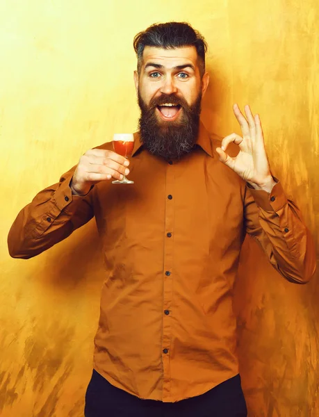Bearded man, long beard. Brutal caucasian smiling happy hipster with moustache in brown shirt holding alcoholic red shot and showing ok on golden texture backgroun