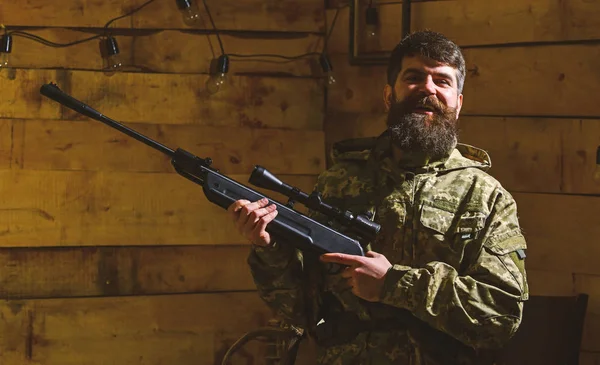 Hunter, brutal hipster with gun prepare rifle for hunting, copy space. Huntsman concept. Man with beard wears camouflage clothing, wooden interior background. Macho on happy face at gamekeepers house — Stock Photo, Image