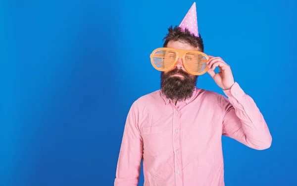 Homem com barba e bigode no rosto calmo solitário em seu aniversário, fundo azul. O tipo de chapéu de festa celebra sozinho. Conceito de aniversário. Hipster em óculos de sol gigantes celebrando aniversário, espaço de cópia — Fotografia de Stock