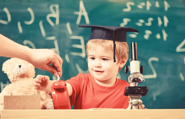 Concepto de descanso escolar. Niño en la cara sonriente mira el despertador. Alumno esperando el receso escolar. Niño en gorra académica cerca del reloj en mano masculina, aula, pizarra de fondo — Foto de Stock