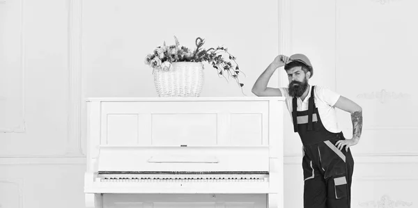 Hombre con barba, trabajador con mono y casco apoyado en el piano, fondo blanco. Courier entrega muebles en caso de mudanza, reubicación. El cargador mueve el instrumento de piano. Concepto de servicio de entrega — Foto de Stock