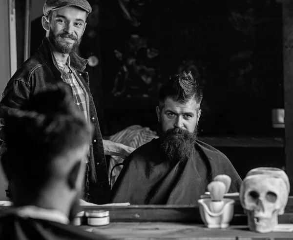 Клиент парикмахера и хипстера с бородой, проверяющий стрижку в зеркале, темный фон. Хипстер и парикмахер разговаривают во время стрижки. Человек с бородой объясняет прическу, которую он предпочитает. Концепция обслуживания — стоковое фото