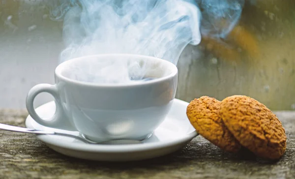 Enjoying coffee on rainy day. Coffee time on rainy day. Fresh brewed coffee in white cup or mug on windowsill. Wet glass window and cup of hot caffeine beverage. Coffee drink with oat cookies dessert