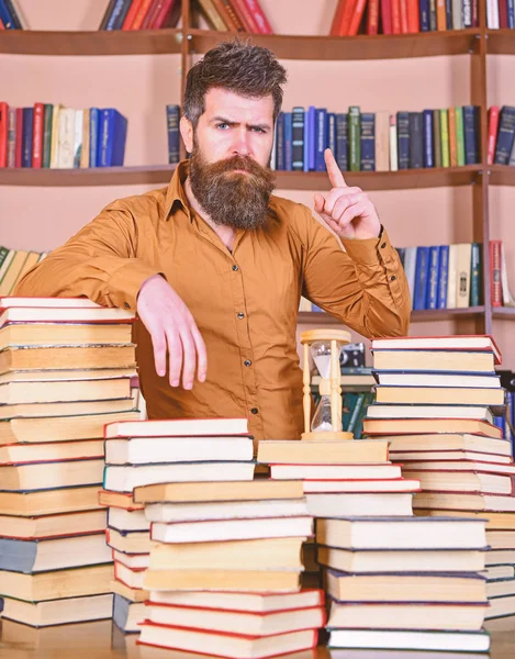 Insegnante o studente con barba si trova a tavola con libri, sfocato. Concetto bibliotecario. L'uomo sulla faccia sicura sta tra mucchi di libri, mentre studia in biblioteca, scaffali su sfondo — Foto Stock