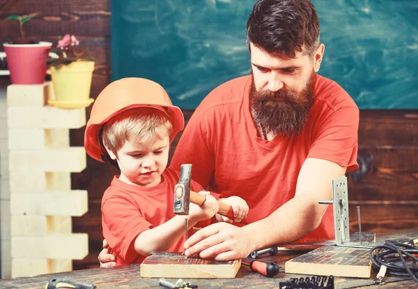 Концепция мастера. Отец с бородой учит маленького сына пользоваться инструментами, молотком, доской на заднем плане. Мальчик, ребенок занят в защитном шлеме, учится пользоваться молотком с папой. — стоковое фото