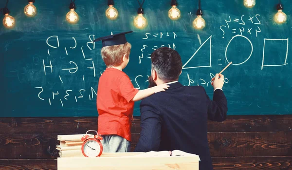 Individual education concept. Teacher and schoolboy turned back in classroom. Kid in graduation cap holding adult male by shoulder. Man pointing with the pen at the shapes drawn on green board — Stock Photo, Image