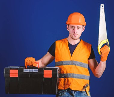 Kask, kask içinde adam araç kutusu taşır ve el testeresi, mavi arka plan tutar. İşçi, repairer, tamirci üzerinde ciddi bir yüz taşıyan araç, onarım, kopya alanı için hazır. Marangoz kavramı