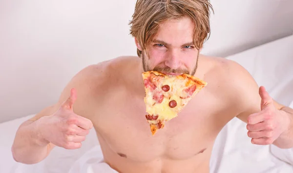 Man likes pizza for breakfast. Who cares about diet. Handsome man holds a piece of pizza in his hands and is about to eat it. Pizza on bed.