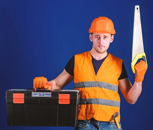 Man in helmet, hard hat carries toolbox and holds handsaw, blue background. Worker, repairer, repairman on serious face carries toolbox, ready for repair, copy space. Carpenter concept — Stock Photo, Image