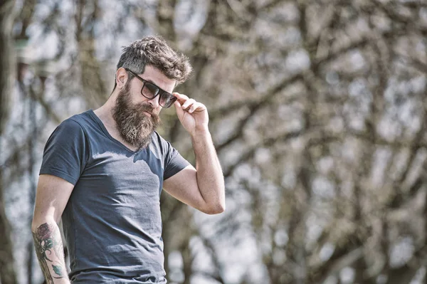 L'homme barbu enlève ses lunettes de soleil le jour ensoleillé. Concept de masculinité. Homme à longue barbe semble élégant et confiant. Homme avec barbe et moustache sur le visage strict, branches sur le fond, déconcentré — Photo