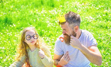 Baba ve kızı, grassplot, arka planı yeşil çimenlerin üzerinde oturur. Aile açık havada eğlence harcamak. Çocuk ve babası crown ve gözlük ile poz booth öznitelikleri fotoğraf. En iyi arkadaşlar kavramı