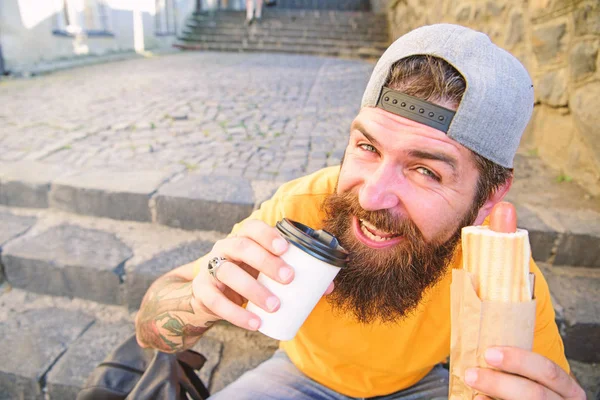 Hipster desfrutar de cachorro quente e beber xícara de papel. Homem barbudo desfrutar de comida de rua fundo urbano. Salsicha saborosa em pão torrado. Conceito de cultura alimentar urbana. Hipster comer cachorro-quente enquanto se sentar em escadas ao ar livre — Fotografia de Stock