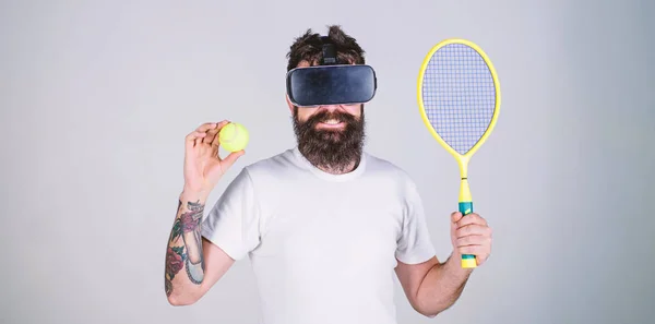 Guy with VR glasses play tennis with racket and ball. Man with beard in VR glasses play tennis, grey background. Hipster on smiling face use modern technology for sport games. Virtual sport concept