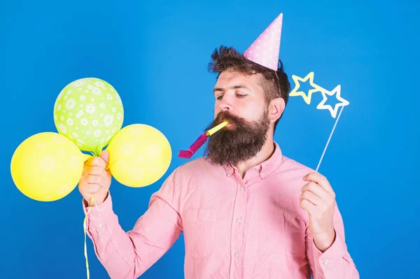 Hipster avec des lunettes en forme d'étoile souffle dans la corne de fête. Homme avec barbe sur le visage calme tient des ballons à air, fond bleu. Concept de surprise. Guy en chapeau de fête avec des attributs de vacances célèbre — Photo