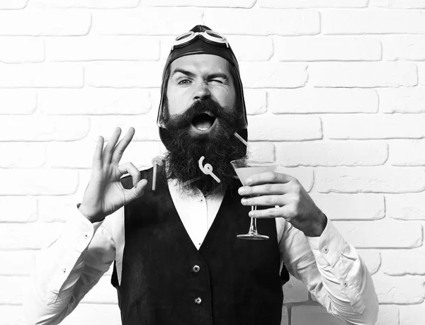 Homem aviador barbudo bonito com barba longa e bigode no rosto engraçado segurando vidro de coquetel alcoólico em jaqueta de couro de camurça vintage com chapéu e óculos no fundo da parede de tijolo branco — Fotografia de Stock