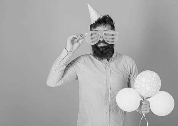 Lächelnder bärtiger Mann mit riesiger Brille, der Spaß am Feiern, am Glücklichsein hat. Mann mit Hipster-Bart und Schnurrbart in rosa Hemd auf blauem Hintergrund. Künstler mit Geburtstagskappe posiert mit Spielzeug-Luftballons — Stockfoto