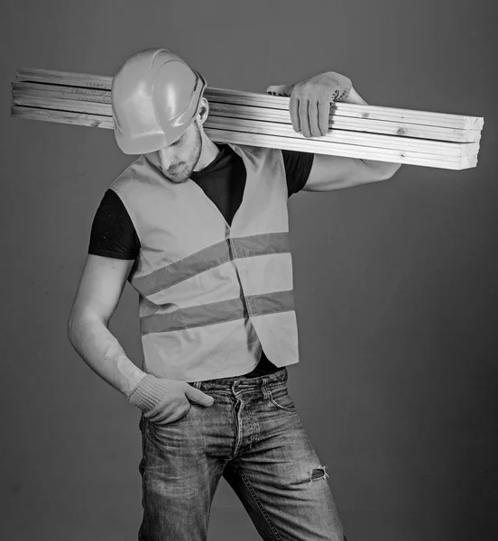 Concepto de trabajador resistente. Hombre con casco, sombrero duro y guantes de protección sostiene la viga de madera, fondo gris. Carpintero, carpintero, obrero, constructor en la cara ocupada lleva vigas de madera en el hombro —  Fotos de Stock