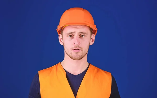 Man in protective helmet, hard hat and working uniform, blue background. Worker, contractor, builder on calm face looking at camera. Protective equipment concept. Builder in helmet posing, copy space