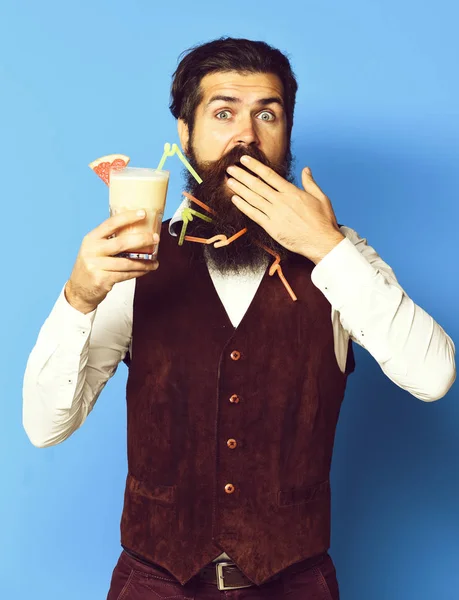 Homem barbudo bonito com barba longa e bigode tem cabelo elegante no rosto surpreso segurando vidro de coquetel alcoólico em jaqueta de couro de camurça vintage no fundo do estúdio azul — Fotografia de Stock