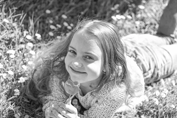 Flicka på leende ansikte innehar röd tulip flower, Njut doften. Ungdom och bekymmerslös koncept. Barn njuta av soliga vårdag liggandes på äng med blommor. Flicka liggande på gräset, grassplot på bakgrund — Stockfoto