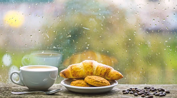 Coffee time on rainy day. Fresh brewed coffee in white cup or mug on windowsill. Wet glass window and cup of hot caffeine beverage. Coffee drink with croissant dessert. Enjoying coffee on rainy day