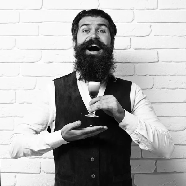 handsome bearded man with long beard and mustache has stylish hair on happy face holding glass of alcoholic shot in vintage suede leather waistcoat on white brick wall studio background