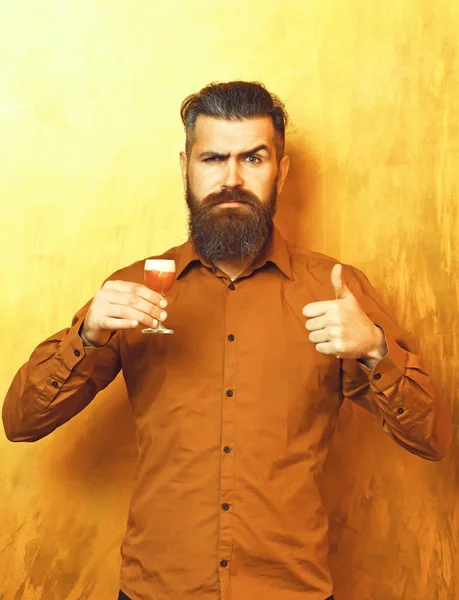 Bearded man, long beard. Brutal caucasian serious hipster with moustache in brown shirt holding alcoholic red shot on golden texture backgroun