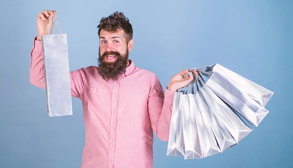 Man with happy face and trendy beard holding shopping bags. Shopaholic spending all salary on clothes. Bearded man in pink shirt isolated on blue background. Man with hipster beard enjoying shopping