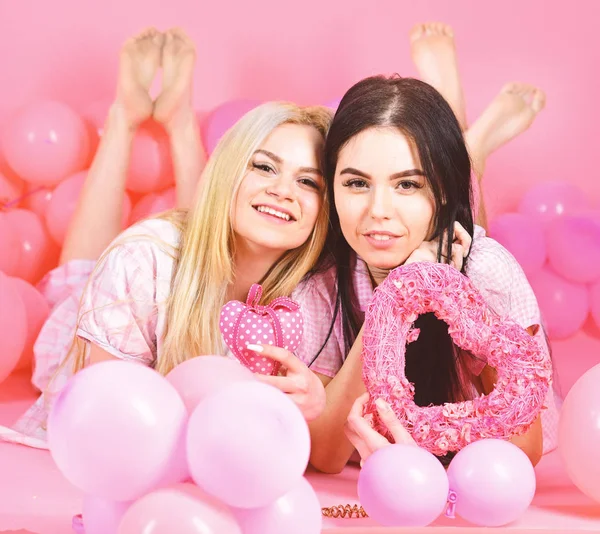 Mujeres en pijama rosa posando en cámara con corazones rosas mientras yacían cerca de globos aéreos en pijama, fiesta de pijamas sobre fondo rosa. Morena y rubia mejores amigos —  Fotos de Stock