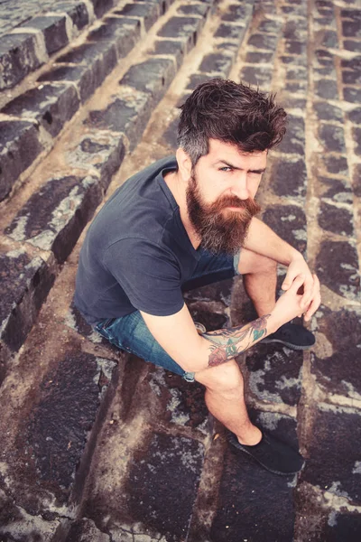 Homme avec barbe et moustache assis sur fond d'escalier en pierre. Hipster sur le visage strict relaxant en plein air. Concept après la pluie. Guy se reposant, profiter de la fraîcheur après la pluie — Photo