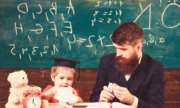 Boy, child on busy face holds crumpled paper in hand while teacher talk to kid. Tries and mistakes concept. Teacher with beard, father teaches little son in classroom, chalkboard on background