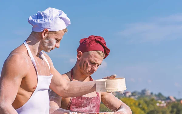 Team of attractive bakers works with flour, preparing dough. Bakers in hats and aprons works with sieve, kneading dough. Brigade of muscular cooks sift flour through sieve. Companions concept