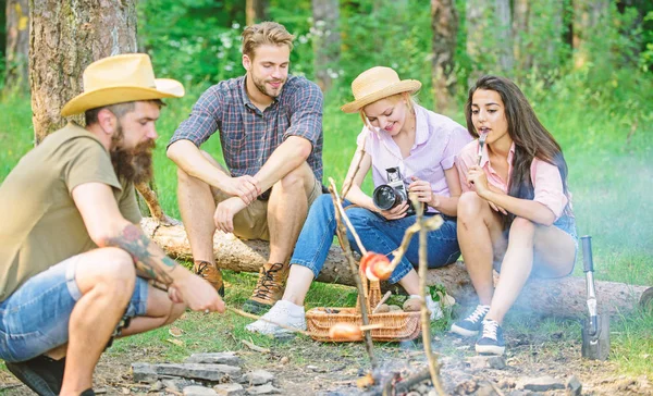 Turistas que têm tempo de lanche com assado sobre comida de fogo. Grupo de amigos turista relaxante perto de fogueira. Hipster assar salsicha enquanto amigos falando impressão de compartilhamento e assistir fotos na câmera — Fotografia de Stock