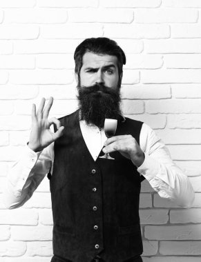Uzun sakallı ve bıyıklı yakışıklı adamın ciddi yüzünde şık saçlar var. Beyaz tuğla duvar arka planında klasik süet deri yelek giymiş alkolik bardakları tutuyor.